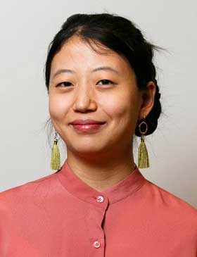 A person with dark hair pulled up in front of a gray background.