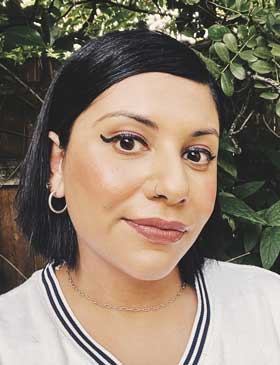 A person with dark hair and hoop earrings in front of a leafy background.
